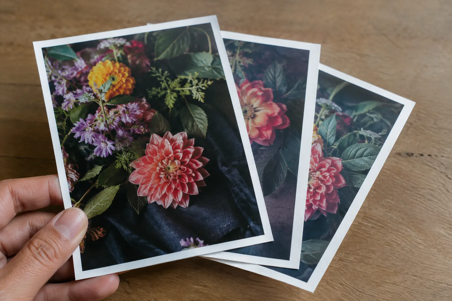 Een hand houdt drie overlappende bloemenprints met witte randen boven een houten tafel